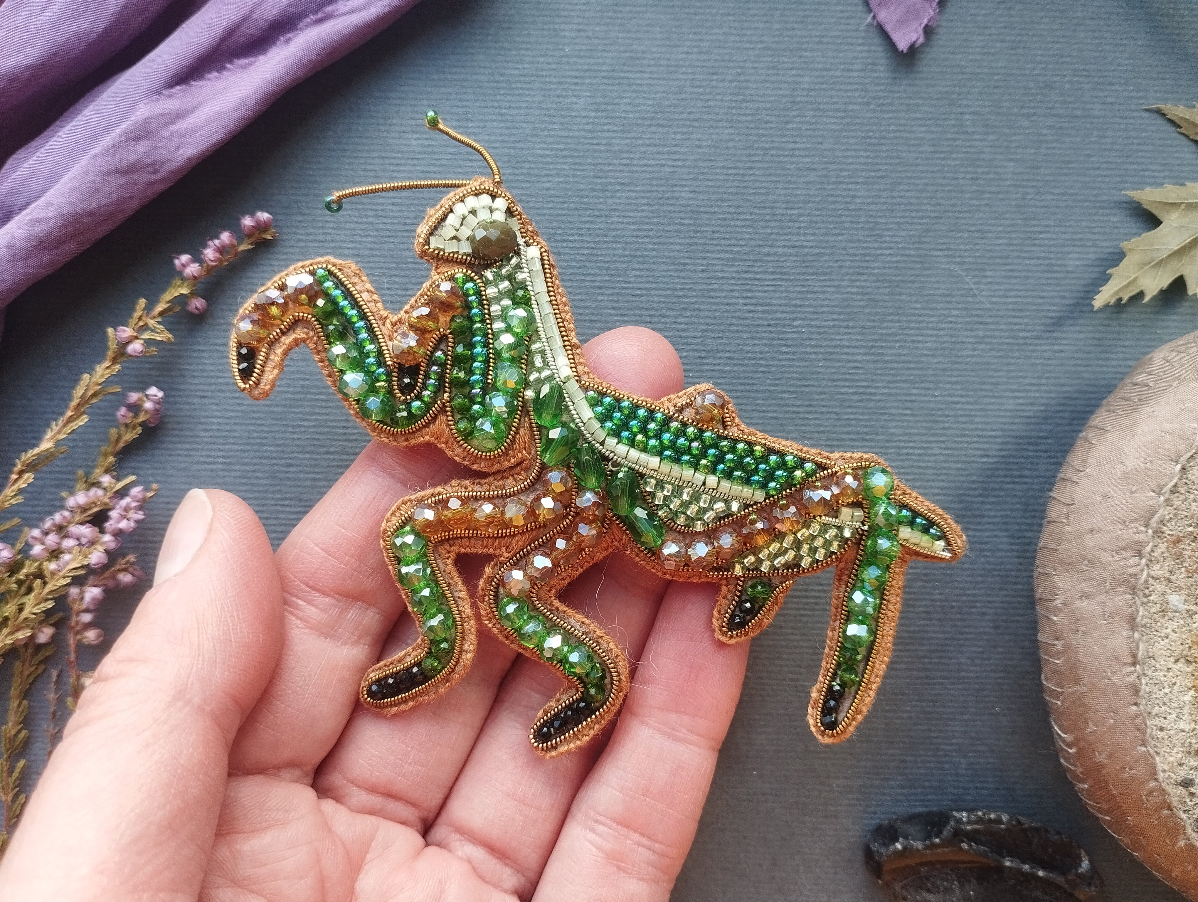 A hand holding a decorative beaded insect, possibly a grasshopper or cricket, against a backdrop of various natural elements such as leaves, flowers, and a stone.